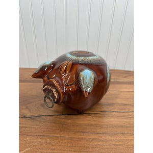 May include: A ceramic piggy bank with a brown glaze and blue accents. The pig has a coin slot on its back and a cork with a metal ring on its snout. The bank is on a wooden surface.