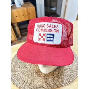Puede incluir: Gorra de camionero roja con un parche blanco con el texto "HUGO SALES COMMISSION" y la marca "PURINA CHOWS". La gorra tiene una parte trasera de malla y una visera curva. La gorra se muestra sobre una superficie tejida.