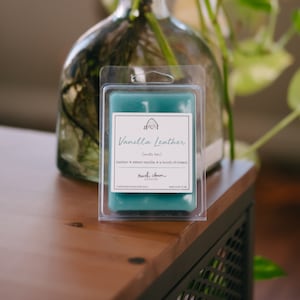 May include: A teal wax melt in a clear plastic clamshell package. The label reads "Vanilla Leather" with scent notes. The wax melt is on a wooden surface, with a glass vase and greenery in the background.