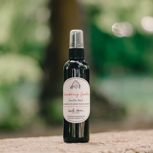 May include: A black glass bottle with a clear spray nozzle. The bottle has a white label with the text "Strawberry Leather (smells like) Strawberry Leather Picnic by the fire" and "rustic charm candles rusticbarmcandies.com room/car spray 4 oz."
