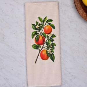 May include: A rectangular waffle-weave kitchen towel in a light cream color featuring a centered botanical illustration of a branch with three bright orange citrus fruits, deep green leaves, and small white blossoms printed on the fabric.
