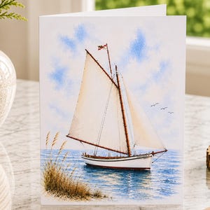 May include: A white greeting card featuring a watercolor illustration of a white sailboat with tan sails on blue water. The scene includes tall golden beach grass in the foreground, a light blue sky with soft clouds, and three small birds in flight.