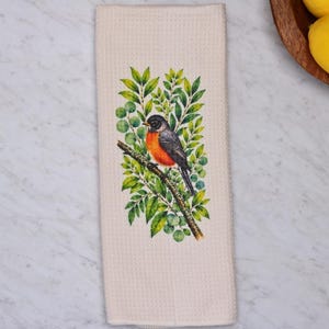 May include: A cream-colored waffle-weave kitchen towel featuring a central illustration of a robin perched on a brown branch surrounded by vibrant green leaves. The bird has a distinct orange breast, a dark head, and a small yellow beak.