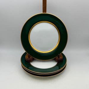 May include: Set of four white ceramic plates with a dark green border and gold trim. The plates are stacked on a wooden stand, showcasing their elegant design. The plates have a classic, refined aesthetic.