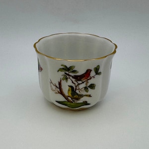 May include: A white porcelain cup with a gold rim and a floral design featuring birds. The cup has a scalloped edge and is decorated with a delicate floral pattern and three birds.