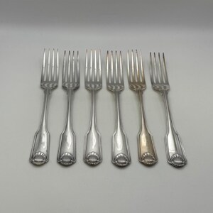 May include: Six silver forks with a shell design on the handles are arranged on a white surface. The forks have tines and a decorative pattern. One fork has a different color than the others.