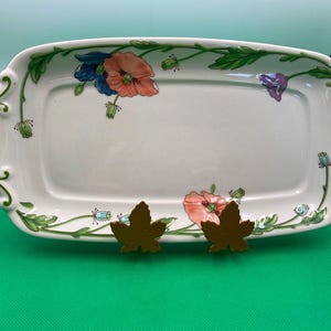 May include: A white ceramic serving dish with a floral design featuring pink, blue, and green poppies. The dish has a scalloped edge and is decorated with green leaves and vines.