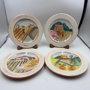 May include: Four decorative ceramic plates with hand-painted rural landscape scenes. Each plate features a different design with fields, houses, and trees, in shades of green, brown, and orange. The plates have a rustic, aged appearance.