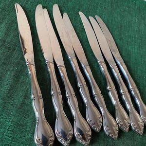 May include: A set of eight silver dinner knives with ornate handles. The knives have a polished finish and are arranged in a row.