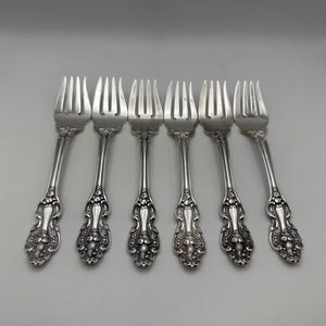 May include: Six silver forks with ornate floral and scrollwork detailing on the handles. The tines are evenly spaced. The forks are arranged in a row against a white background, suggesting a formal dining setting.