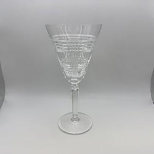 May include: A clear crystal wine glass with a detailed, etched design on the bowl. The glass has a long stem and a round base. The bowl features a series of horizontal and vertical cuts, creating a textured appearance.