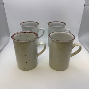 May include: Four ceramic mugs with a speckled, light-tan finish and a brown rim. Each mug has a curved handle and a cylindrical shape. The mugs are arranged in a square formation against a white background.