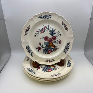 May include: A stack of four white ceramic plates with scalloped edges. Each plate features a floral design with pink, blue, and orange accents. The plates are stacked on a wooden plate stand.
