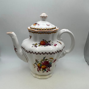 May include: White ceramic teapot with a lid, decorated with colorful fruit and floral patterns. The teapot has a curved spout, a large handle, and a decorative finial on the lid. The design includes oranges, grapes, and other fruits.