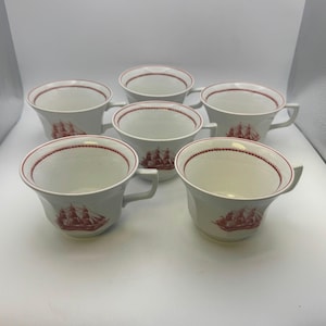 May include: Six white teacups with a red ship design. Each cup has a handle and a red trim around the rim. The ship design is repeated on each cup, creating a cohesive set.
