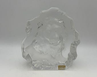 May include: A clear glass sculpture of a lion's head and upper body. The lion is etched into the glass, with a frosted appearance. The sculpture has an irregular, organic shape. A small, rectangular label is at the base.