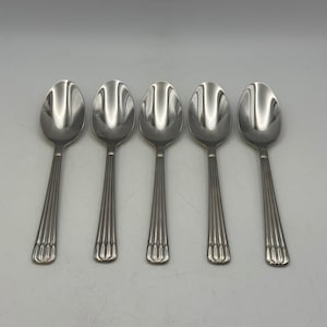 May include: Five stainless steel spoons arranged in a row. Each spoon has a rounded bowl and a handle with vertical grooves. The spoons are made of a shiny, silver-colored metal and are set against a plain, light gray background.