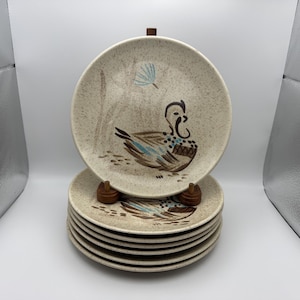 May include: A stack of seven round ceramic plates with a speckled beige finish. Each plate features a hand-painted illustration of a duck in brown, blue, and black tones, accompanied by a stylized blue floral element and brown grass markings.
