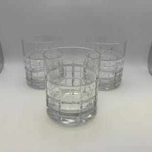 May include: Three clear, patterned drinking glasses. The glasses are cylindrical with a grid-like design etched into the glass. The glasses are empty and sit on a white surface.