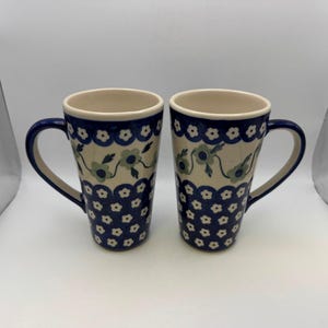 May include: Two ceramic mugs with a blue and white floral pattern. The mugs have a cream-colored interior and a dark blue handle. The exterior features a repeating pattern of blue flowers and a scalloped border. The mugs are tall and cylindrical.