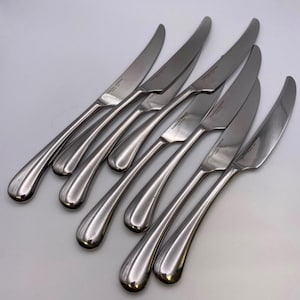 May include: A set of eight silver dinner knives with curved handles. The knives have a polished finish and are arranged in a fan shape.