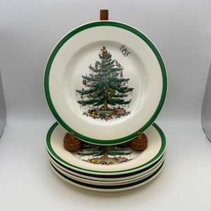 May include: A stack of white ceramic plates with a green border. Each plate features a Christmas tree design in the center. The plates are stacked on a wooden stand, showcasing a festive holiday theme.