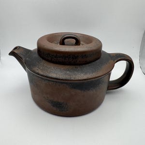 May include: A brown ceramic teapot with a matte finish and dark charcoal-colored mottled accents. The teapot features a cylindrical body, a curved handle, a short spout, and a circular lid with a small central handle on top.