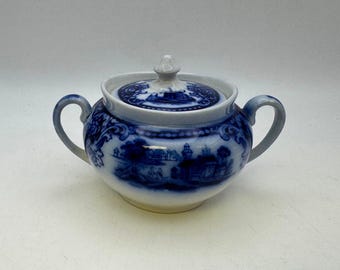 May include: A blue and white ceramic sugar bowl with a lid. The bowl features a detailed landscape design and two handles. The lid has a small knob for easy removal. The sugar bowl is a classic piece of tableware.