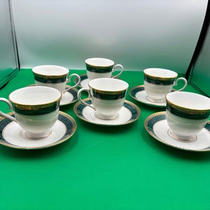 May include: Set of six white teacups and saucers with a green and gold floral pattern. The cups and saucers are arranged in a circle on a green surface.