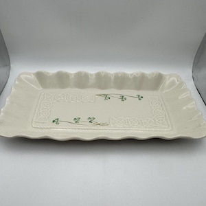 May include: A rectangular off-white ceramic serving tray with a scalloped edge. The center features an embossed Celtic knot border and two small green shamrock illustrations positioned diagonally across the flat surface of the dish.