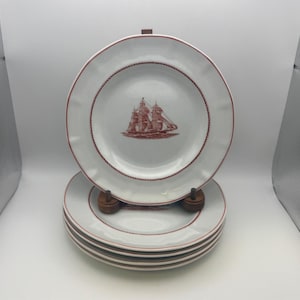 May include: A set of white ceramic plates with a red ship illustration in the center. The plates have a red border and are stacked on a wooden plate stand. The plates are a classic design.