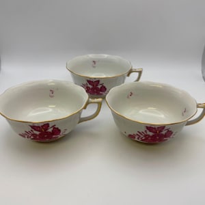 May include: Three white teacups with gold trim and handles, each adorned with a red floral pattern. The cups have a scalloped rim and are arranged on a white surface, showcasing a classic design.