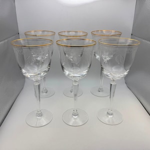 May include: Set of six clear wine glasses with gold rims. Each glass features a floral etched design and a long stem. The glasses are arranged in a row, showcasing their elegant shape and delicate details, perfect for a special occasion.