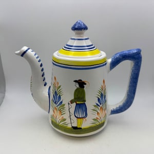 May include: A white ceramic teapot with a blue handle and spout, and a lid with blue and white stripes. The teapot features a painted design of a person in traditional clothing and floral accents. The teapot is approximately 8 inches tall.
