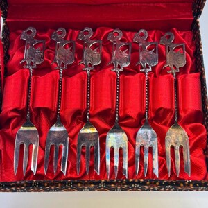 May include: A set of seven silver forks with decorative tops, presented in a red velvet-lined box. The forks have a detailed design on the handles and are arranged neatly within the box, suggesting a vintage or collectible item.