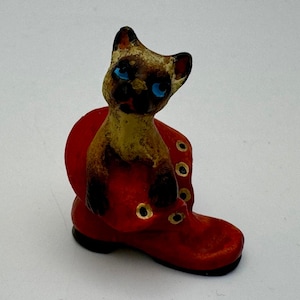 May include: A vintage figurine of a Siamese cat with blue eyes, perched inside a red boot. The cat has tan and brown fur with black paws and ears. The boot has black soles and gold eyelets.