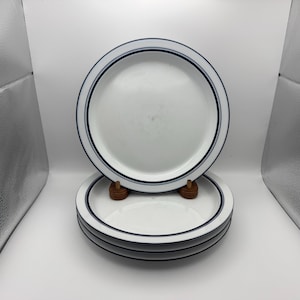 May include: A stack of three white ceramic plates featuring a simple design with two thin, dark blue circular bands running along the outer rim. The plates are round with a smooth finish and a clean, minimalist aesthetic suitable for dining.