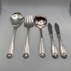 May include: A set of silver-colored serving utensils, including a spoon, fork, ladle, and two butter knives. Each piece has a decorative shell design at the base of the handle. The utensils are arranged on a light gray surface.