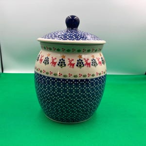 May include: A ceramic jar with a lid, featuring a blue and white pattern. The jar has a festive design with reindeer and Christmas trees. The lid is topped with a dark blue knob. The jar is approximately 6 inches tall.
