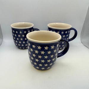 May include: Three ceramic mugs with a navy blue background and a pattern of white stars. The mugs have a cream-colored interior and dark blue handles. The mugs are arranged on a white surface.