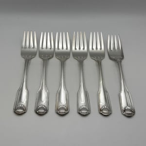 May include: Six silver forks arranged on a light gray surface. Each fork has a decorative shell design at the base of the handle. The tines are evenly spaced, and the forks are clean and polished.
