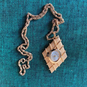 May include: A gold chain necklace with a gold-toned watch pendant. The watch face is white with black markings and hands. The pendant is shaped like a diamond.