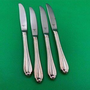 May include: Four silver dinner knives with ornate handles. The knives have a polished finish and are arranged in a row on a green surface.