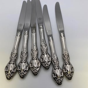 May include: Six silver-plated dinner knives with ornate handles. The knives have a floral design and the words "18/8 Stainless" are engraved on the blades.