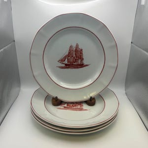 May include: Set of four white ceramic plates with a red border and a red illustration of a sailboat. The plates have a scalloped edge and are stacked on top of each other.