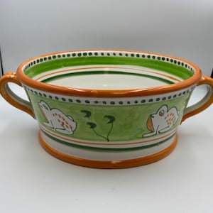 May include: A white ceramic serving bowl with two handles. The bowl is decorated with green and orange stripes and a green background with two white frogs and green leaves.