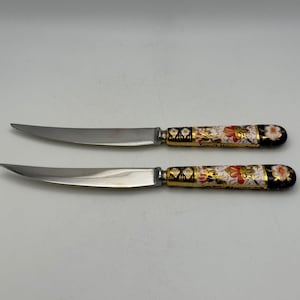 May include: Two stainless steel knives with ornate handles. The handles feature a white base with floral designs in red, gold, and black. The blades are curved and pointed, suitable for a dining setting.