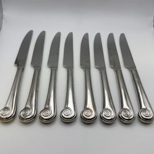 May include: A set of eight silver-colored knives. Each knife has a curved blade and a handle with a spiral design at the end. The knives are arranged in a row on a white surface.