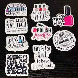 May include: A collection of nail art stickers with various phrases and designs. The stickers feature text like "Life is Prettier with Polish," "But First, Nails," and "Nail Tech." The color palette includes pink, black, white, and blue.