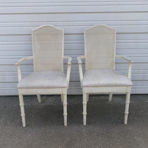 Puede incluir: Un par de sillones de color blanco roto con respaldos altos y arqueados con paneles de rejilla trenzada. Cada silla tiene un asiento acolchado tapizado en tela clara y cuatro patas con un diseño segmentado similar al bambú.